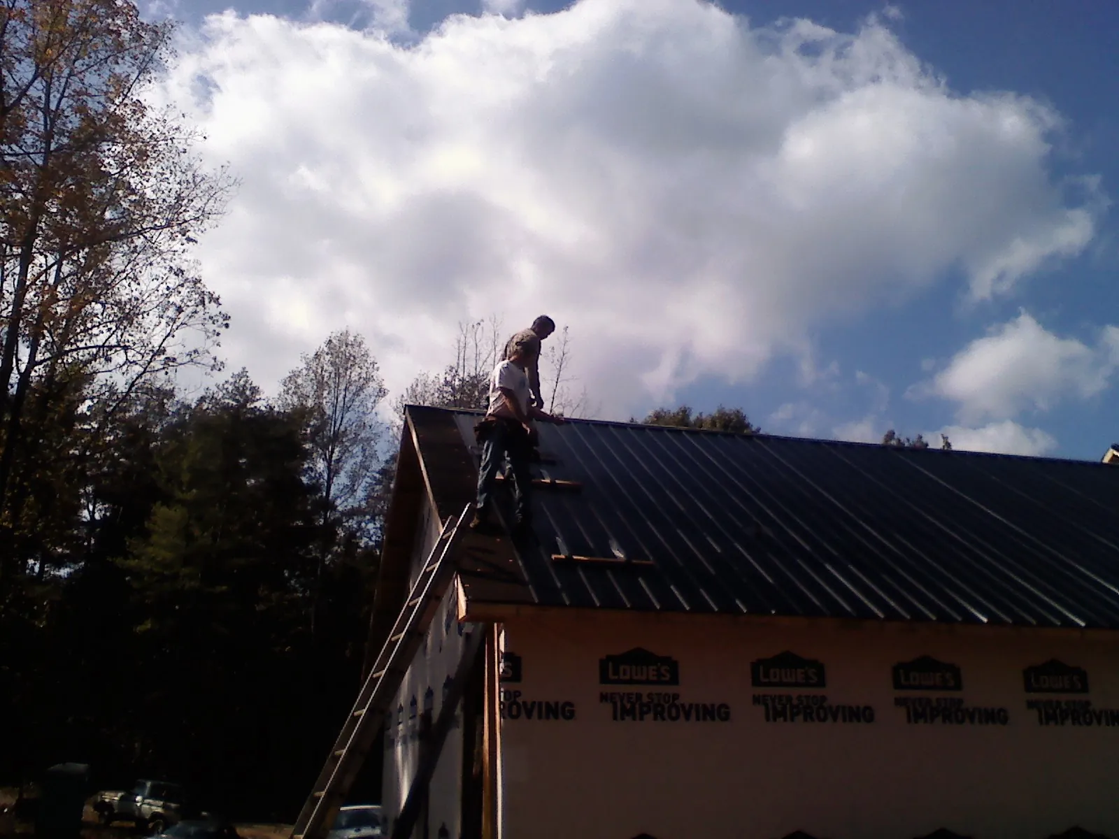 Roofer fastening dark metal panels high against a cloudy sky, from a Roof Inspection job in Newfield